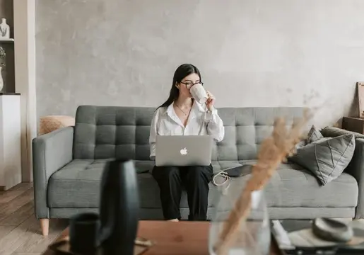 young woman sitting on couch drinking out of a mug while working on apple mac