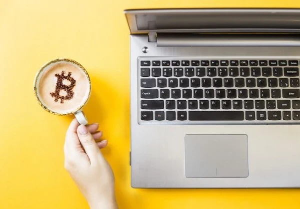 Coffee mug with Bitcoin latte art next to an Macbook laptop