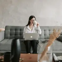 young woman sitting on couch drinking out of a mug while working on apple mac