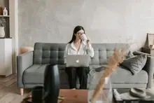 young woman sitting on couch drinking out of a mug while working on apple mac